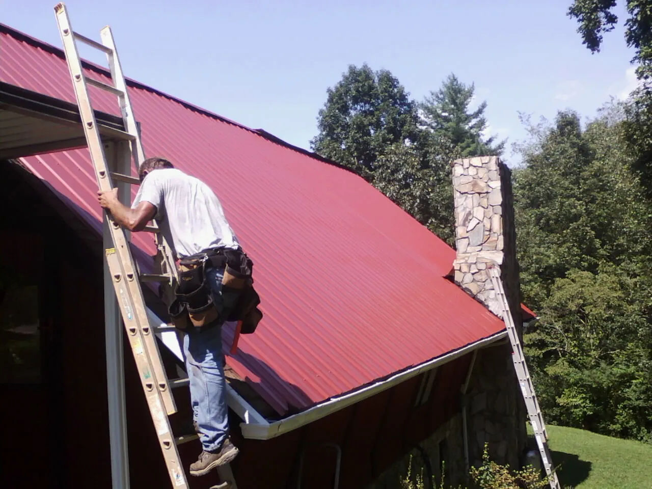 Roofer on a ladder beside a red metal roof and stone chimney — Soffit & Fascia Repair by Roofing Brothers in Myrtle Creek